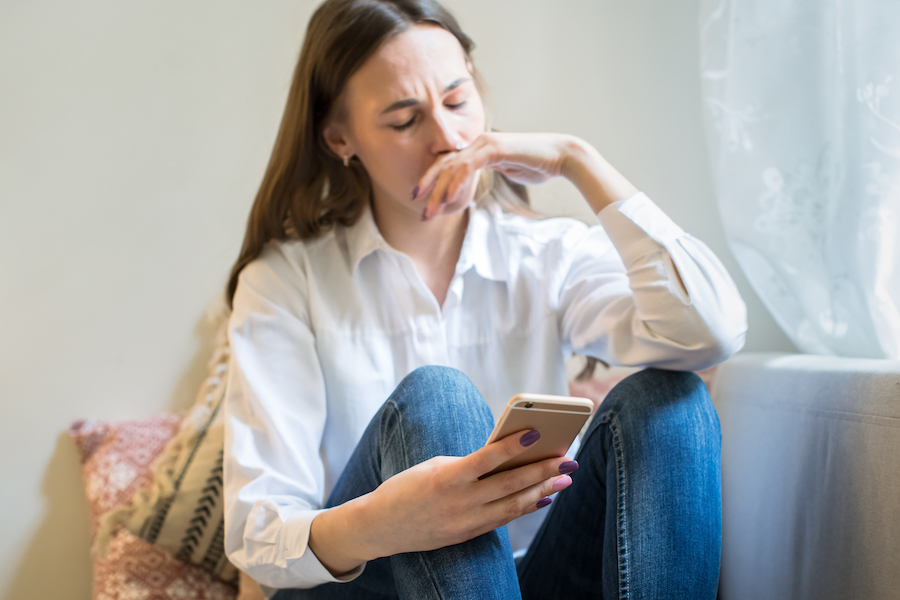 Crying young woman in depression looking at phone gets bad news, covering mouth with hand Young woman anxiously looking at phone